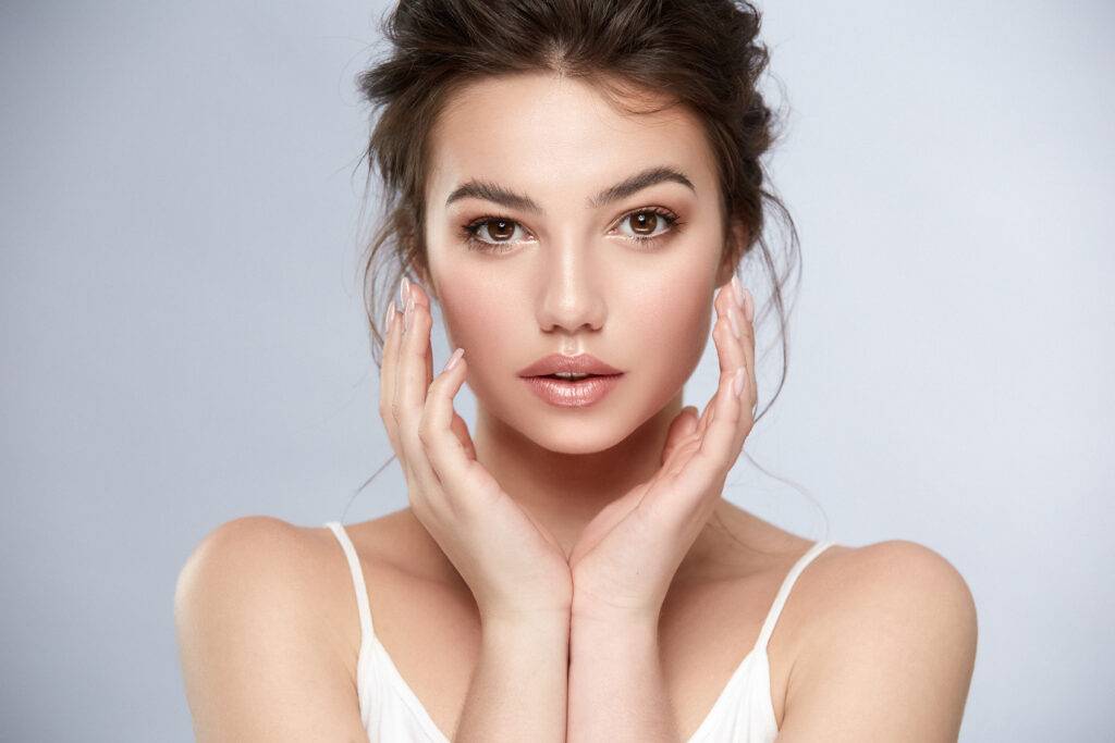 Portrait of a woman with natural makeup, flawless skin, and neatly styled hair, posing with her hands gently framing her face against a soft gray background