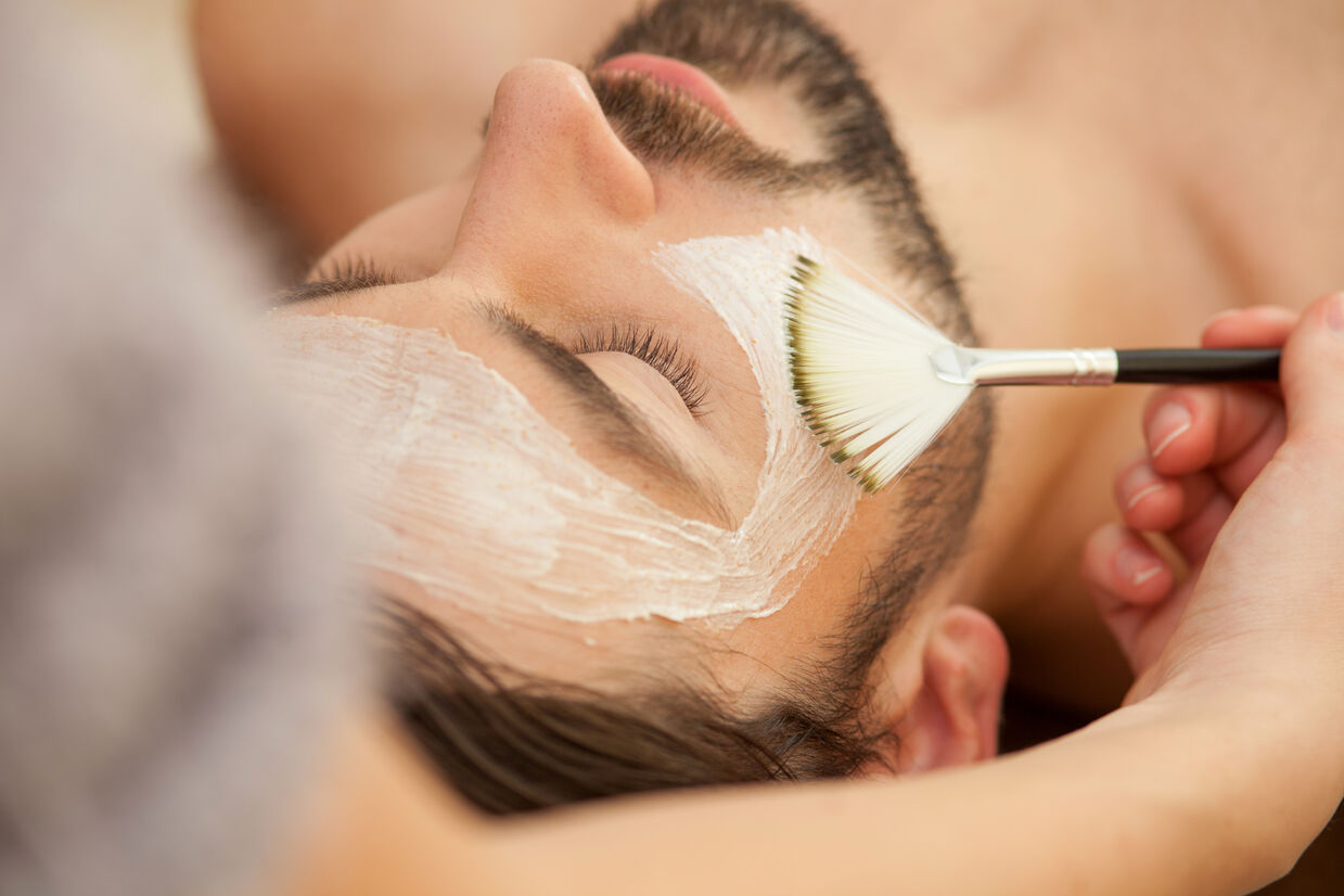 Close-up of a man receiving a facial treatment, with a beauty professional applying a white mask to his face using a fan brush, creating a relaxing spa experience.