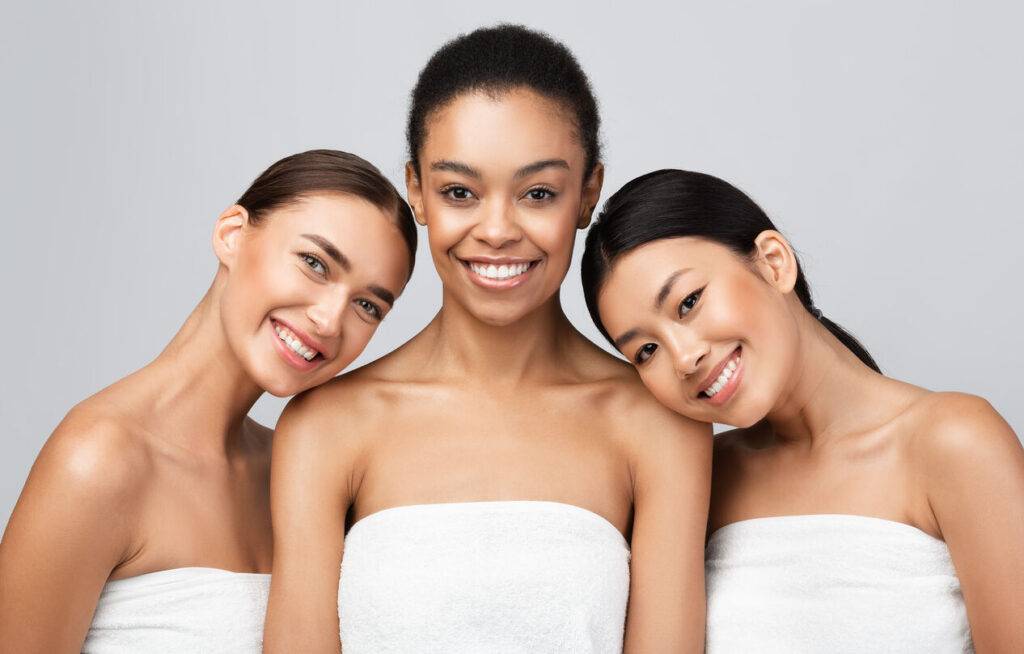 Portrait of three diverse women with glowing skin, smiling confidently and wearing white spa towels, symbolizing beauty and self-care, against a light gray background.
